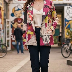 Puede incluir: Una chaqueta de manga larga con un estampado gráfico multicolor de personajes de Mickey Mouse en tonos rojos, amarillos y negros. La chaqueta tiene solapas de muesca, dos bolsillos delanteros y se lleva sobre una camiseta de tirantes color crema.