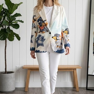 Könnte beinhalten: Ein cremefarbener Blazer mit einem bunten Grafikdruck von Dackeln in verschiedenen Outfits, kombiniert mit einem schlichten weißen T-Shirt, einer weißen Hose und weißen Turnschuhen. Der Blazer hat einen Knopfverschluss und ein steigendes Revers.