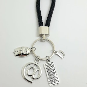 Può includere: Un portachiavi in ​​pelle nera con un anello portachiavi argentato e ciondoli. I ciondoli includono un mouse per computer, un simbolo chiocciola, una tastiera e la lettera D.
