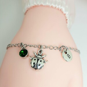 Può includere: Un braccialetto a catena d'argento con tre ciondoli: una pietra preziosa verde, una coccinella d'argento e un ciondolo d'argento con la lettera "B" su di esso.