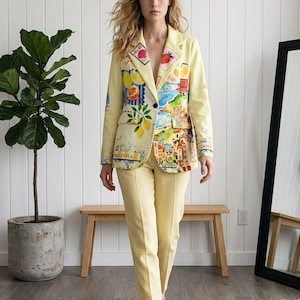 May include: A pale yellow blazer and matching trousers set. The blazer features a single-button closure, notched lapels, and vibrant, multi-colored graphic patches depicting lemons, strawberries, coastal landscapes, and the text DOLCE VITA.