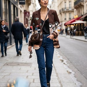 May include: A brown blazer featuring various graphic patches and prints, worn over a black camisole with blue denim jeans. The outfit includes a brown leather crossbody bag, gold layered necklaces, and black loafers with gold chain hardware.