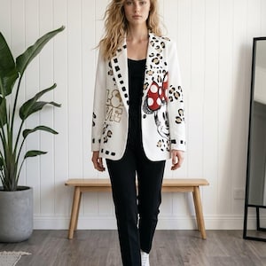 Könnte beinhalten: Ein weißer Blazer mit einem schwarz-braunen Leopardenmuster, einer Grafik einer Zeichentrickfigur mit einer roten, gepunkteten Schleife und dem Wort LOVE in Blockbuchstaben. Der Blazer wird über einem schwarzen Oberteil und einer schwarzen Hose mit weißen Turnschuhen getragen.