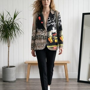 Op de afbeelding: Een blazer met een zwart en beige kleurverloop, witte graffiti-tekst, rode hartaccenten en een afbeelding van Mickey Mouse. Het jasje wordt gecombineerd met een effen zwarte broek en witte sneakers voor een complete outfit.