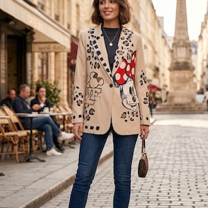 Könnte beinhalten: Ein beigefarbener Blazer mit einem Grafikdruck von Minnie Maus, schwarzen Leopardenflecken und geometrischen Formen, getragen über einem dunklen Oberteil mit blauer Jeans, weißen Turnschuhen und einer kleinen braunen Handtasche. Der Blazer hat einen Knopf.