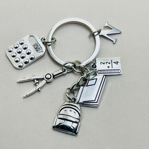 Può includere: Portachiavi argentato con una calcolatrice, una bussola, un quaderno, uno zaino e un ciondolo a forma di lettera W.