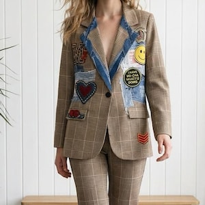 Könnte beinhalten: Ein brauner Blazer mit Karomuster und dazu passender Hose, verziert mit Denim-Revers und verschiedenen bunten Aufnähern, darunter ein gelber Smiley, ein rotes Herz und ein runder Aufnäher mit dem Text I HAVE NO ORA WHATEU DOMS.