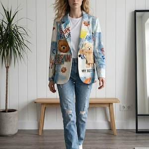 Pode incluir: Um blazer de ganga de lavagem clara com texto colorido estilo graffiti, motivos de sol pintados e dois apliques de ursos de peluche nos painéis frontais. O blazer é usado sobre uma t-shirt branca e calças de ganga azuis desgastadas.