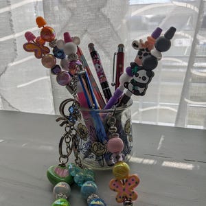 May include: A glass jar filled with colorful pens and keychains. The keychains feature beads, butterflies, and animal charms. The pens have colorful designs and some have animal toppers.