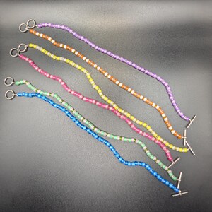 May include: Six colorful beaded eyeglass chains in purple, orange, yellow, pink, blue, and green. Each chain has a silver metal loop at one end and a T-bar clasp at the other end. The beads are small and round.