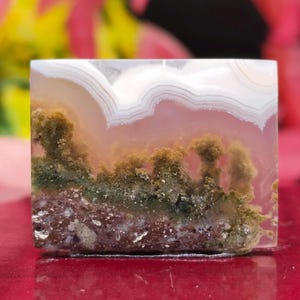 May include: A square-cut, polished gemstone with a landscape-like pattern. The top features white and tan bands, transitioning to a pinkish-purple midsection. The bottom displays a forest-like design with green and brown hues, creating a natural, artistic appearance.