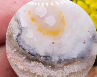 以下が含まれることがあります： ユニークな模様の丸く磨かれた宝石。石は、グレー、黒、オレンジの渦巻き模様の白いベースが特徴です。下端はテクスチャ加工された結晶の外観をしています。宝石は人が持っています。