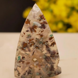 May include: A triangular-shaped polished gemstone featuring a translucent, milky white base with scattered inclusions of metallic copper-brown, dark brown, and small flecks of teal green mineral deposits throughout the stone's surface.