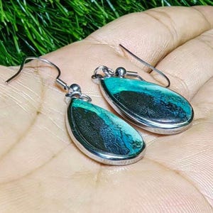 May include: A pair of teardrop-shaped earrings with a silver-colored metal frame. The earrings feature a striking combination of teal, turquoise, and black colors. The earrings are displayed on a hand, showcasing their design and craftsmanship.