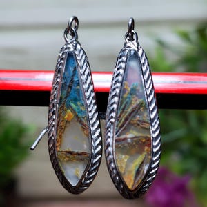 May include: A pair of teardrop-shaped earrings with a silver-colored, textured frame. The earrings feature a colorful, abstract design with shades of blue, yellow, and brown. The earrings are hanging against a blurred background.
