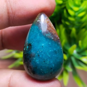 May include: A teardrop-shaped gemstone with a polished surface. The stone features a striking blend of colors, including deep teal, brown, and hints of black. The gemstone is held between two fingers, showcasing its size and shape.