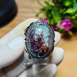 May include: A silver ring with an oval-shaped gemstone. The gemstone features a mix of colors, including red, green, and white, with a clear, glossy finish. The ring has a detailed, textured setting.
