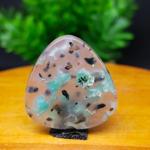 May include: A polished, triangular gemstone with a mix of colors. The stone features a base of peach and tan hues, speckled with black and brown spots. Patches of light green and teal add contrast. The gemstone is mounted on a small, black base.