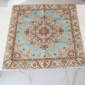 May include: A square scarf featuring a central medallion design in shades of light blue, orange, black, and cream. The fabric displays an intricate paisley pattern throughout the center and a decorative border with floral and geometric motifs.