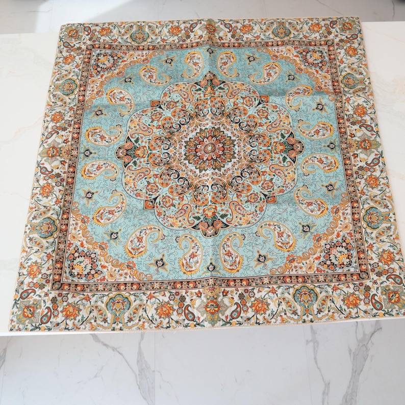 May include: A square scarf featuring a central medallion design in shades of light blue, orange, black, and cream. The fabric displays an intricate paisley pattern throughout the center and a decorative border with floral and geometric motifs.