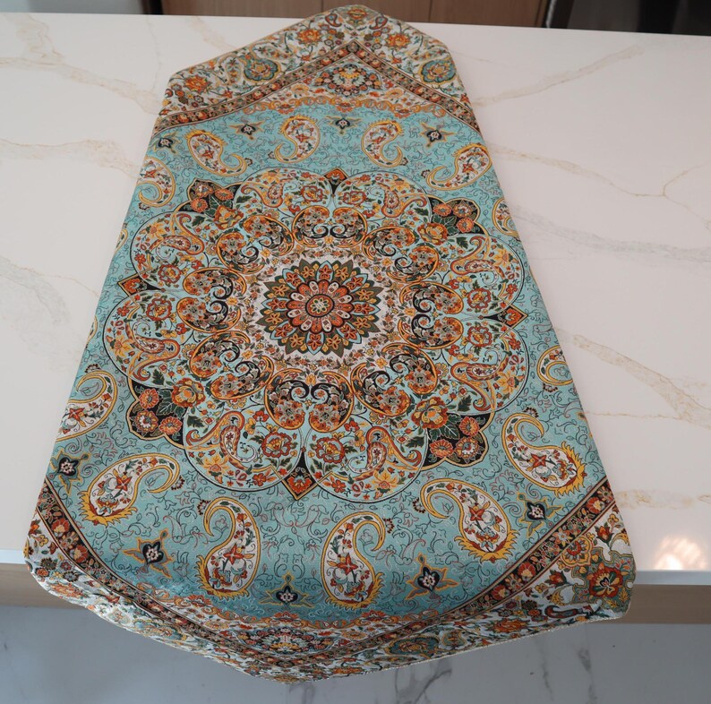 May include: A rectangular table runner featuring a light blue background with a central circular mandala pattern. The design includes intricate paisley motifs, floral elements, and geometric shapes in shades of orange, black, and white.