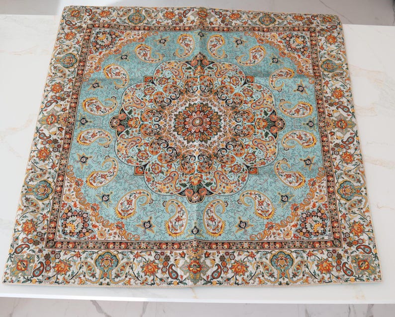 May include: A square textile featuring a central medallion and intricate paisley patterns in shades of light blue, orange, black, and cream. The design includes a detailed border with repeating floral motifs and teardrop shapes throughout the fabric.