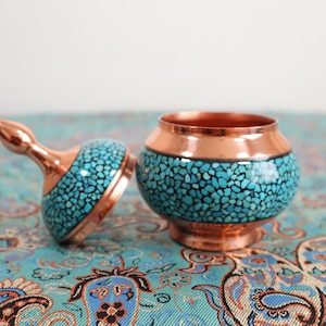 Puede incluir: Un tarro decorativo de cobre y turquesa con tapa. El tarro está decorado con pequeñas piedras turquesas que están engastadas en el cobre.