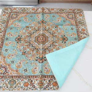 May include: A square fabric mat featuring a light blue background with a central medallion and intricate paisley patterns in orange, black, and white. The mat has a solid light blue underside that is partially folded over to reveal the plain backing.
