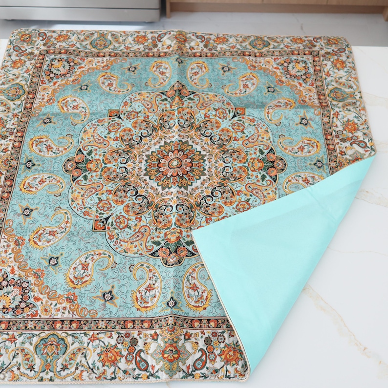 May include: A square fabric mat featuring a light blue background with a central medallion and intricate paisley patterns in orange, black, and white. The mat has a solid light blue underside that is partially folded over to reveal the plain backing.