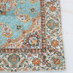 May include: A square textile featuring a light blue central panel with intricate orange, yellow, and dark green paisley and floral motifs. A wide border displays a dense pattern of colorful flowers and paisley shapes on a white background.