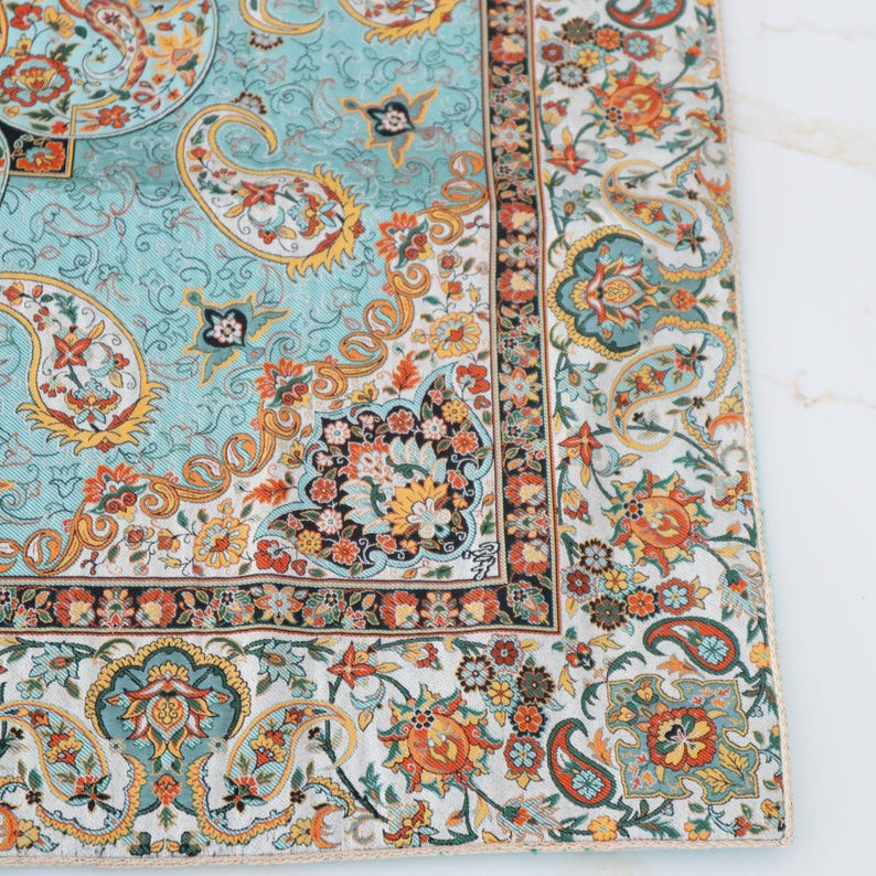 May include: A square textile featuring a light blue central panel with intricate orange, yellow, and dark green paisley and floral motifs. A wide border displays a dense pattern of colorful flowers and paisley shapes on a white background.