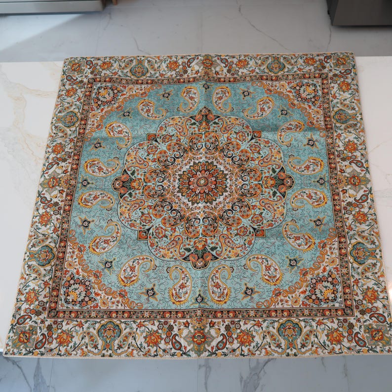 May include: A square fabric scarf featuring a central medallion design with intricate paisley and floral patterns in shades of turquoise, orange, black, and cream. The border displays a repeating motif of paisley shapes and floral elements.
