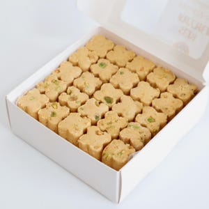 May include: A white box filled with flower-shaped, light brown sweets. Some of the treats have green flecks, likely pistachios, adding color and texture. The box is open, revealing the treats inside.
