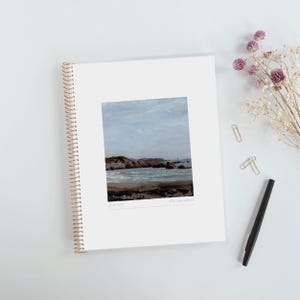 May include: A white spiral-bound notebook featuring a centered rectangular painting of a coastal landscape with a sandy beach, dark rocks, and a light blue sky. The text 2020-2027 and Miss Sara Atkins is printed in small grey font below the artwork.
