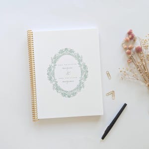 May include: A white spiral-bound planner with a gold wire coil. The cover features a sage green ornate oval frame surrounding the text "TWO THOUSAND twenty-six & TWO THOUSAND twenty-seven" in a serif font. The planner is accompanied by a black pen.