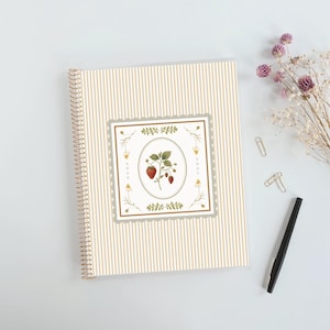 May include: A spiral-bound notebook with a beige and white vertical striped cover. The center features a square frame with a scalloped edge, containing an illustration of a strawberry plant inside an oval border, with the text 2026 and 2027.