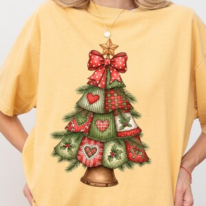 Puede incluir: Camiseta de color mostaza con un diseño de árbol de Navidad. El árbol está decorado con lazos rojos, corazones y patrones de cuadros. Una estrella dorada corona el árbol y una campana forma la base. La palabra "Mustard" está impresa abajo a la izquierda.