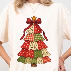 Puede incluir: Camiseta color marfil con un diseño de árbol de Navidad. El árbol está compuesto por secciones de patchwork en rojo, verde y crema, adornado con un lazo rojo y una estrella dorada. La palabra "Ivory" está impresa en la parte inferior.
