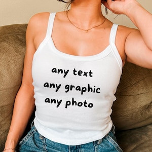 May include: White tank top with the text "any text any graphic any photo" in black. The top has a scoop neck and spaghetti straps. The person is wearing blue jeans.