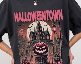 Puede incluir: Camiseta negra con un diseño de Halloween. El diseño presenta el texto rosa "Halloweentown", una casa embrujada, una calabaza tallada y el texto "EST. 1996". La camiseta está siendo usada.