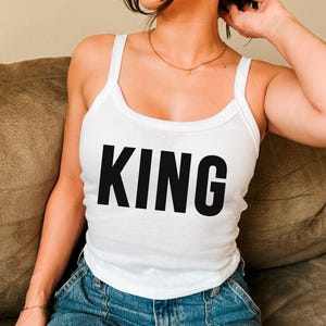 Könnte beinhalten: Weißes Tanktop mit dem Wort "KING" in fetten schwarzen Buchstaben aufgedruckt. Das Oberteil hat einen Rundhalsausschnitt und Spaghettiträger. Die Person trägt eine blaue Jeans.