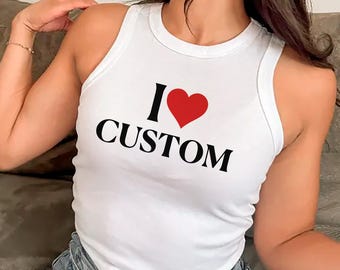 May include: White tank top with the text "I ♥ CUSTOM" in black and a red heart. The top is sleeveless and has a round neckline. The model is wearing blue jeans.
