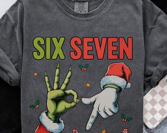May include: Dark gray sweatshirt with the words "SIX SEVEN" in green and red. The design features a green hand making an "OK" sign and a white hand pointing, both wearing Santa hats. Candy canes and stars are also included.