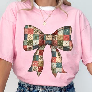 Könnte beinhalten: Hellrosa T-Shirt mit einem großen Schleifendesign. Der Bogen besteht aus Patchwork-Quadraten in verschiedenen Farben, darunter Rot, Grün und Blau. Der Text "Pink / Comfort colors" befindet sich unten links.