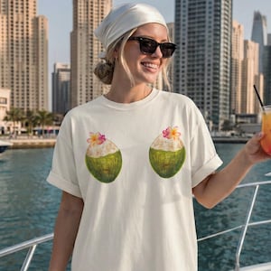 May include: Ivory-colored t-shirt featuring a whimsical design of two coconut halves with flowers. The shirt is a relaxed fit, perfect for a casual, summery look. The shirt is made of a soft, comfortable material.