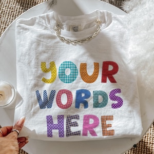 May include: A white short-sleeve t-shirt featuring the text YOUR WORDS HERE in colorful, patterned block letters. Each letter displays a unique design, including polka dots, gingham, and solid colors. A silver chain is attached to the neckline.