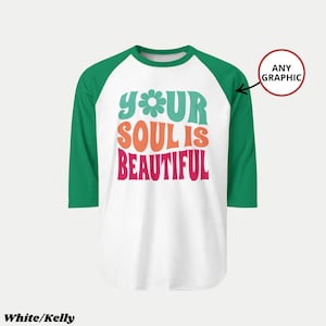 Pu&ograve; includere: Una maglietta da baseball bianca con maniche verdi. La maglietta presenta la scritta "YOUR SOUL IS BEAUTIFUL" in turchese, arancione e rosa, con una grafica floreale. La maglietta &egrave; etichettata "White/Kelly".