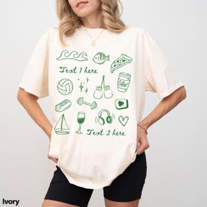 May include: Ivory-colored t-shirt with green doodle illustrations of waves, a fish, pizza, a volleyball, boxing gloves, a coffee cup, a hotdog, a sailboat, a wine glass, headphones, and a heart. The text "Text 1 here" and "Text 2 here" are also included.