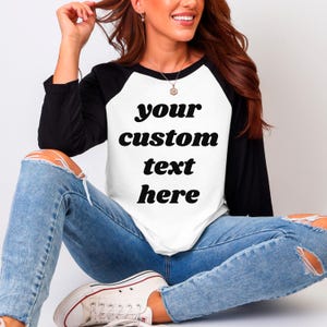 Pu&ograve; includere: Una maglietta raglan bianca e nera con la scritta "your custom text here" in grassetto, in nero. La maglietta ha maniche nere e un corpo bianco. La persona indossa jeans blu e scarpe da ginnastica bianche.