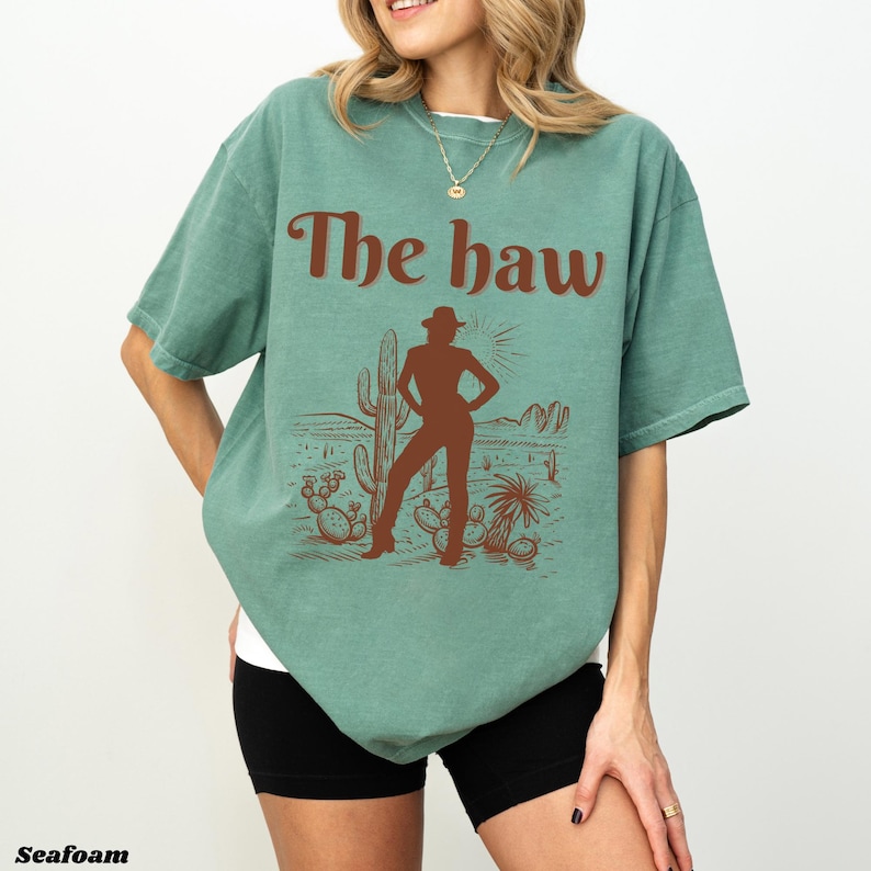 Op de afbeelding: Zeegroen T-shirt met een bruine afbeelding van een woestijnlandschap en de tekst "The haw". De afbeelding toont een persoon met een cowboyhoed, cactussen en een zon. Het shirt heeft een relaxte pasvorm.